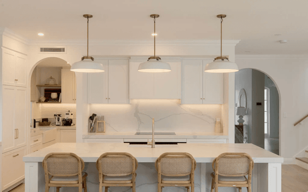 Bright, refined white kitchen centred on a large island with four woven-seat bar stools. The room features full-height white shaker-style cabinetry, a pale marble-look veined splashback, integrated appliances, a light benchtop, brass-toned pendant lights, and an arched opening leading toward the adjacent circulation space.
