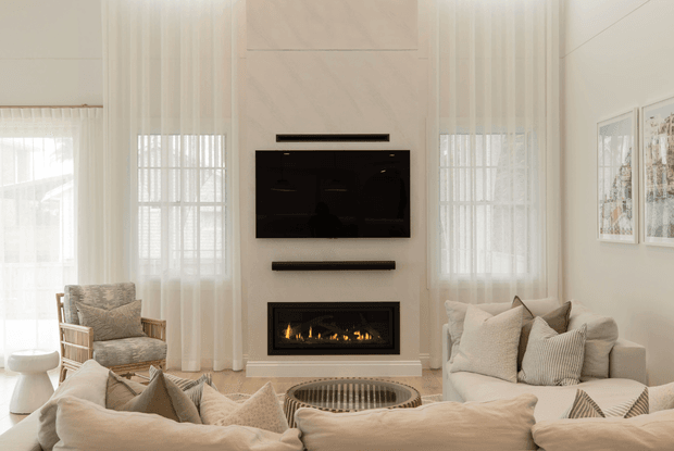 Bright, refined living room with a tall feature wall centred on a wall-mounted black television and linear fireplace. Sheer floor-to-ceiling curtains frame large windows, while layered cream and beige sofas, an upholstered timber-framed armchair, cushions, and a round coffee table create a soft, inviting seating area. Framed artwork and restrained contemporary detailing complete the space.