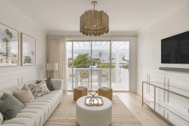 Bright, refined living room with a large curved upholstered sofa, round upholstered coffee table, textured rug, wall-mounted television, slim console, decorative wall panelling, framed artwork, and a timber outdoor deck visible through full-height glazed doors. A sculptural timber pendant adds a statement focal point above the seating area.
