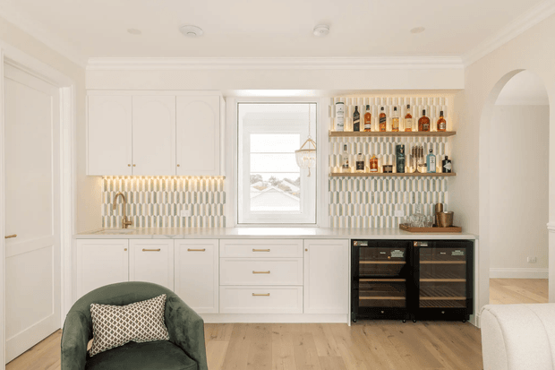 Bright, refined butler's pantry or entertaining kitchenette with white built-in cabinetry, brass hardware, a central window, patterned vertical-tile backsplash, open liquor display shelves, and two black wine refrigerators. The room has pale timber flooring, curved archway detailing, and a green upholstered accent chair in the foreground.