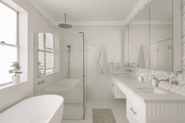 Bright, spacious contemporary bathroom with a freestanding white bathtub, frameless glass walk-in shower, double vanity with white fluted cabinetry, large mirrored wall cabinets, pale tiled walls and floor, and a subtle patterned tile splashback. Natural light enters through a large window, creating a clean, airy atmosphere.