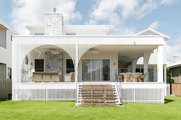 Bright exterior view of a white two-storey coastal-style house with a broad covered front veranda. The facade features extensive vertical white picket-style screening and balustrades, arched openings, white weatherboard cladding, a prominent irregular stone chimney, and central stairs leading from the lawn to the veranda. Outdoor seating is visible beneath the veranda, with neighbouring houses at either side.