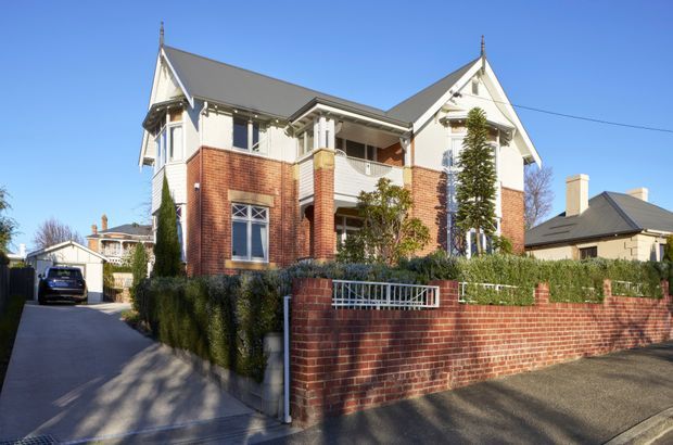 Street-level exterior view of a substantial two-storey brick residence with prominent gabled roof forms, white rendered upper facade details, decorative verandah and balcony elements, and a landscaped front garden enclosed by a red brick boundary wall. A driveway and detached garage are visible to the side, with neighbouring homes in the background.