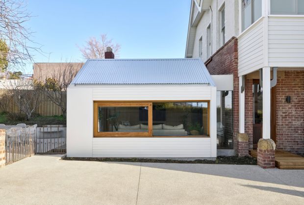 Exterior view of a compact, contemporary glazed pavilion or extension beside a larger brick-and-weatherboard house. The pavilion has white horizontal cladding, a pale corrugated metal gable roof, and a large timber-framed picture window revealing a small furnished interior. A concrete-paved area occupies the foreground, with brick columns and a covered entry or veranda at right.