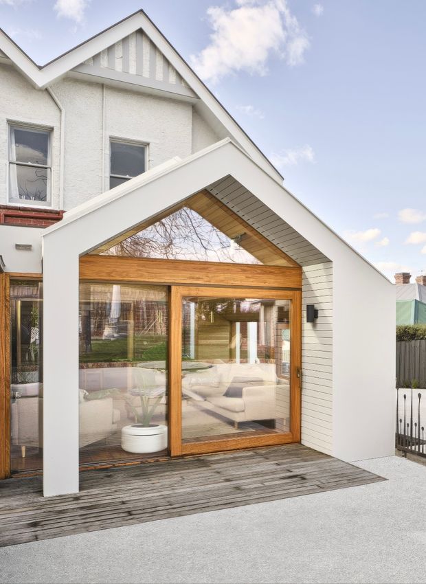 Exterior view of a residential rear extension attached to a traditional gabled house. The extension has a prominent white angular gable-like portal, warm timber-framed full-height glazing, a timber-lined soffit, white horizontal cladding, and a weathered timber deck. A living room is visible through the glazing.