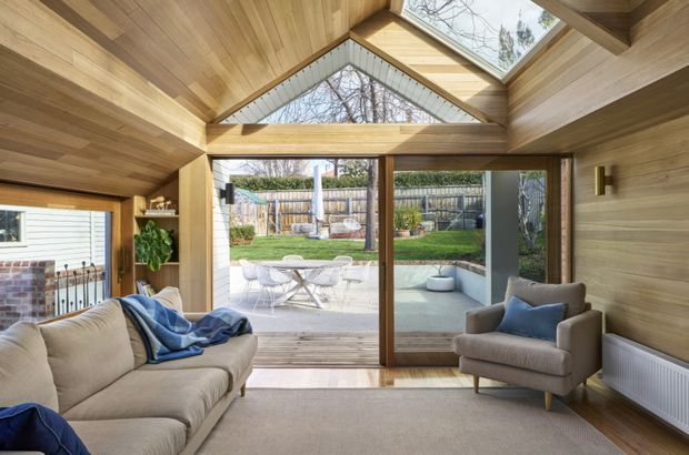 A warm, timber-lined living room opening through large sliding glass doors to a paved patio and landscaped backyard. The room has a pitched, timber-clad ceiling with exposed structural beams, a high triangular glazed gable, and a roof skylight. A neutral sofa and armchair with blue cushions sit on a light carpet, while a brick feature wall and garden are visible beyond the glazing.