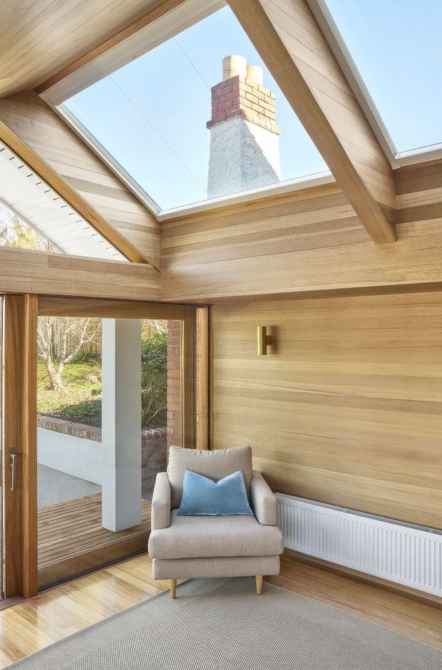 Bright, compact interior sitting area with extensive warm timber lining on the walls and sloped ceiling. A large roof skylight frames a brick chimney, while exposed timber beams and clerestory-like glazing bring in abundant daylight. A glass door opens to an exterior courtyard or garden, and the room contains a light upholstered armchair, blue cushion, radiator, wall sconce, and neutral rug.