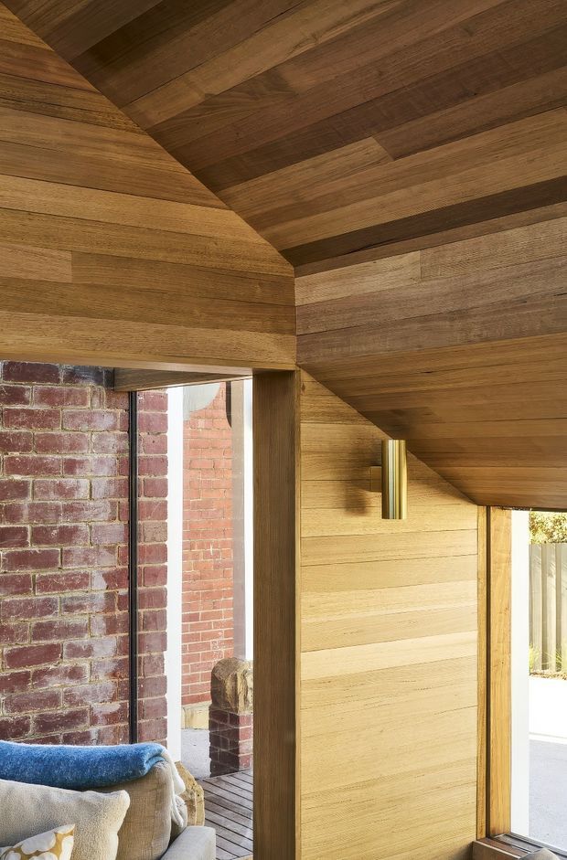 Interior architectural detail showing a warm timber-lined room beneath a sloping ceiling, with matching horizontal timber wall panelling and exposed dark timber beams. A brass cylindrical wall light, large glazed openings, and an adjacent exposed red-brick wall are visible. Part of a sofa appears in the lower left, suggesting a living area. The image emphasizes materiality, joinery, and the transition between timber and brick.