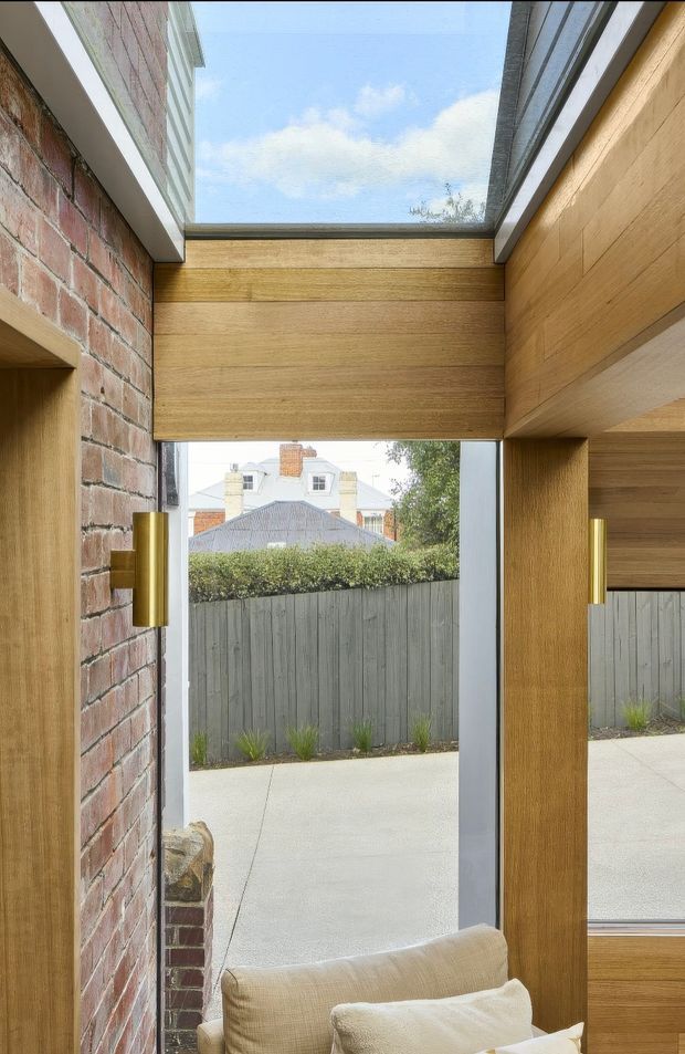 A narrow, light-filled interior space framed by exposed red brick and warm timber wall and ceiling panels. A large central glazed opening and overhead skylight bring in views of the courtyard, fence, neighbouring homes, and sky. Brass wall lights flank the opening, while the lower foreground includes part of a light upholstered sofa. The image highlights a contemporary architectural transition between interior and exterior, with strong natural materials and integrated glazing.