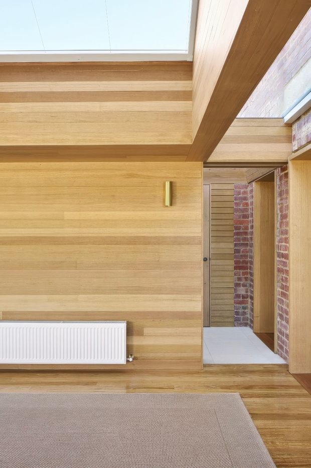Interior view of a warm, timber-lined room opening into a tiled passage or adjoining space. Horizontal timber wall panelling continues across the ceiling and built-in doorway surrounds, with exposed reddish brickwork at right, a large skylight, a slim brass wall light, white radiator, and pale carpet over timber flooring. The composition emphasises natural materials, strong horizontal lines, and integrated architectural detailing.