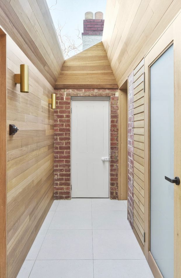 A narrow, bright passageway or entry corridor with pale timber wall and ceiling panelling, white tiled flooring, and exposed red brick framing a white door at the end. A glazed timber-framed door or window lines the right side, while simple cylindrical brass wall lights and dark hardware provide understated accents. The open roof area above reveals a brick chimney, adding a strong material contrast and architectural detail.