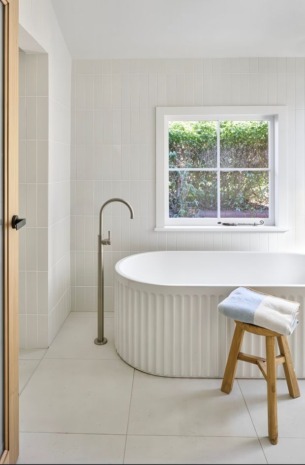 Bright, serene bathroom featuring a freestanding white fluted bathtub beneath a white-framed divided-pane window, pale large-format floor tiles, vertically arranged light wall tiles, and a brushed metal floor-mounted bath tap. A timber stool with a folded towel sits beside the tub, and a light timber door is visible at left.