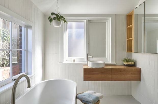 A bright, compact bathroom featuring a freestanding white bathtub beside large windows, white vertical wall panelling, a wall-mounted timber vanity with an integrated white basin, mirrored cabinetry, and open timber shelving. A hanging plant and small stool add soft natural accents. The room has abundant natural light and a clean contemporary feel.