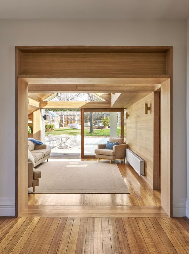 View through a broad timber-lined doorway into a bright living room. The room features warm timber wall and ceiling finishes, polished timber flooring, a neutral sofa and armchair, a large light rug, and extensive glazing opening toward a small garden courtyard. A roof skylight and exposed timber framing bring in natural light.