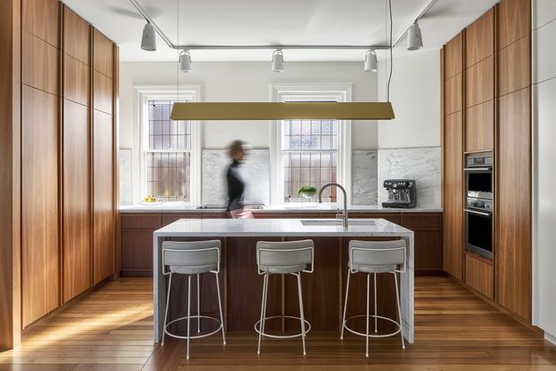 A refined galley-style kitchen with full-height warm timber cabinetry on both sides, a central island with seating for three, and a white stone benchtop. Tall sash windows bring in natural light, while a marble-patterned splashback, integrated stainless-steel appliances, pendant lighting, and timber flooring complete the space. The room has a balanced, contemporary character with strong symmetry and clean built-in detailing.