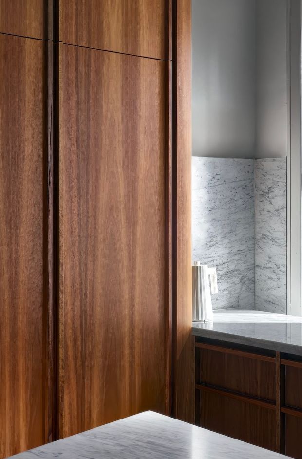 A close-up view of a refined bathroom interior featuring floor-to-ceiling dark timber cabinetry and panelling, a pale veined marble vanity countertop and backsplash, and a contemporary wall sconce. The composition emphasizes the contrast between warm wood grain and cool grey-white stone, with clean-lined built-in joinery and a softly lit, minimalist atmosphere.