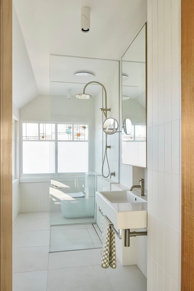 Bright, narrow contemporary bathroom with white tiled walls and floor, a freestanding bathtub behind a frameless glass shower enclosure, and a wall-mounted white vanity with integrated basin. A large mirrored cabinet reflects the shower and stained-glass-style frosted windows. Brass-toned shower and basin fittings, round mirrors, recessed ceiling lights, and timber door or wall framing add warmth to the predominantly white space.