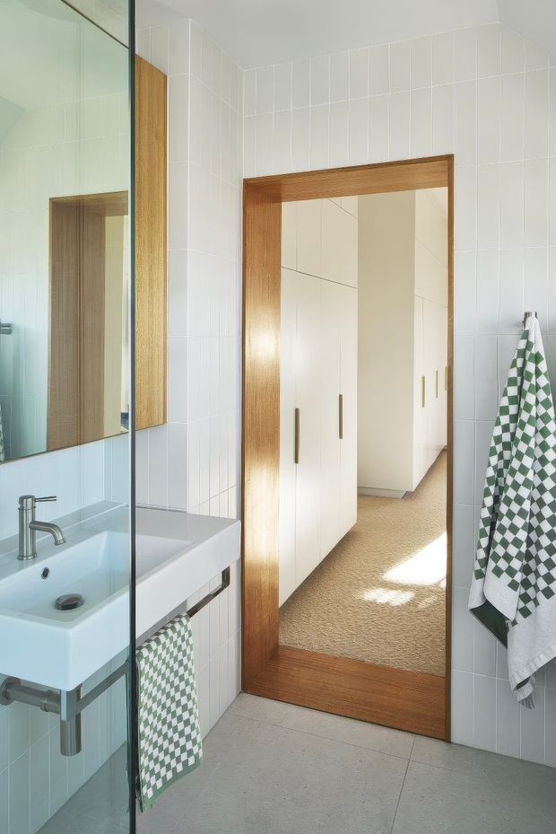 Bright contemporary bathroom with white tiled walls, a wall-mounted white basin, chrome tapware, a clear glass shower screen, and a large timber-framed mirrored cabinet or doorway. A checkered green-and-white towel adds the main accent. The mirror reflects white built-in cabinetry and a carpeted adjoining space.