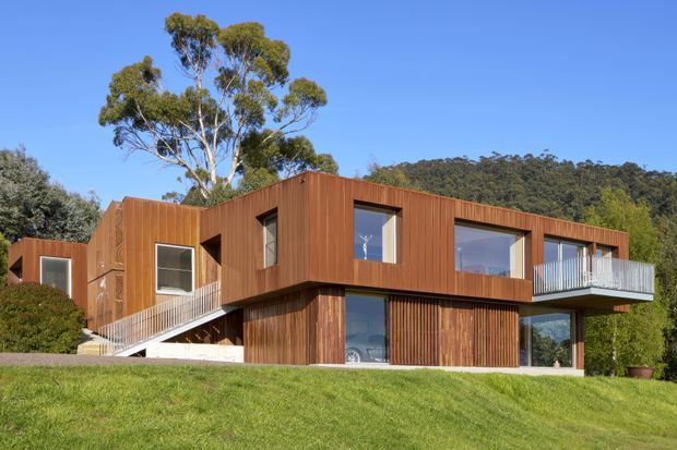 Exterior view of a contemporary multi-level house set on a sloping grassy site, with extensive vertical timber cladding, projecting box-like upper volumes, large windows, a glazed lower level, and a balcony with a metal balustrade. Mature trees and a wooded hillside form the background.