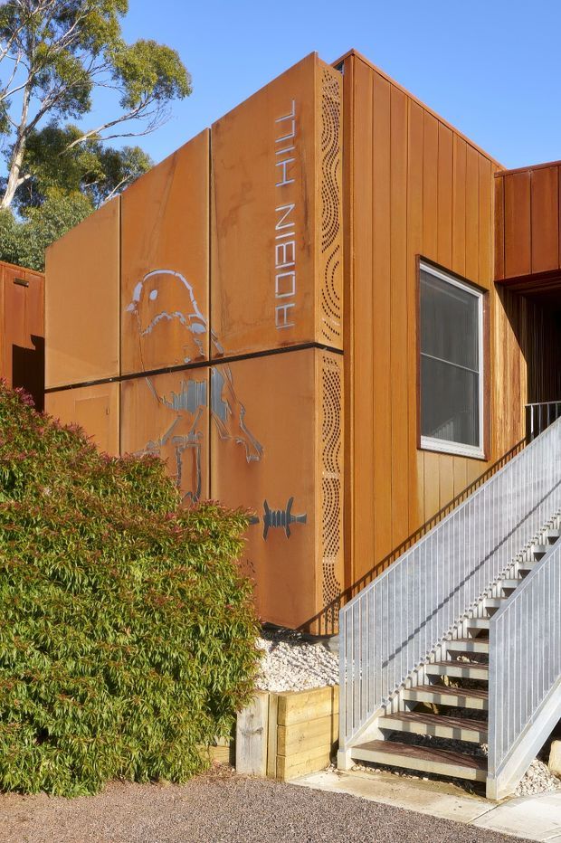 Exterior view of a contemporary building clad in rust-toned weathering steel, with vertical panel joints, perforated decorative screening, and engraved mural-like graphics. A metal staircase with silver balustrades leads to the entrance, while landscaped shrubs partially screen the lower facade.