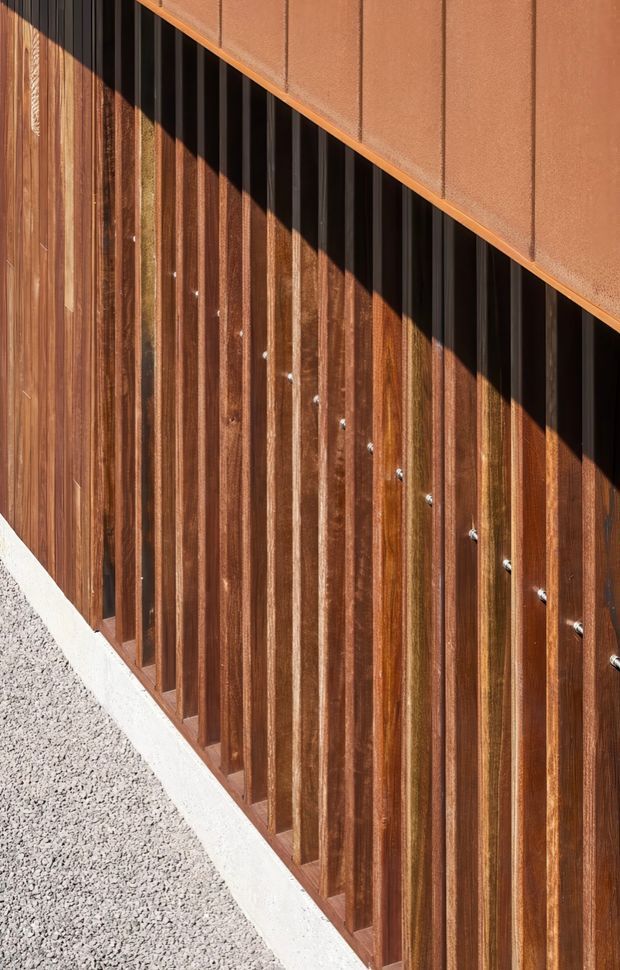 Close-up exterior architectural detail showing vertical timber battens or slats fixed over a dark recessed facade, beneath a diagonally sloping rust-toned metal panel. A pale concrete base and light gravel are visible at the lower edge. Strong sunlight creates a bold triangular shadow across the timber screen.