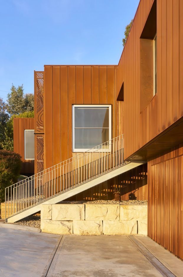 Exterior view of a contemporary multi-level residence clad in warm rust-orange vertical metal panels. The facade features projecting volumes, large rectangular windows, decorative perforated metal screening, and an elevated accessible ramp with slim metal railings. Pale rough-cut stone retaining walls and concrete paving form the foreground, while trees and blue sky sit in the background.