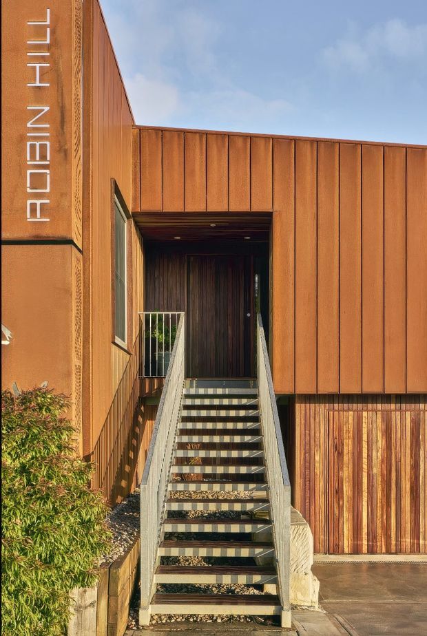 Exterior architectural photograph of a contemporary building entrance, featuring a straight external stairway leading to a recessed dark timber doorway. The facade is clad in warm rust-toned vertical metal panels with timber accents, patterned detailing, and a vertical illuminated sign reading “ROBIN HILL.” Gravel landscaping, planting, and a paved entry area frame the stairs.