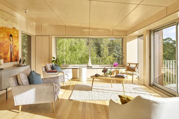 A bright, warm living room with extensive light timber wall and ceiling panelling, wide timber flooring, and large windows and sliding glass doors overlooking leafy greenery. The room contains upholstered lounge chairs and sofas, a low timber coffee table, a small dining or reading setting, artwork, and indoor plants. A balcony or terrace is visible beyond the glazing.