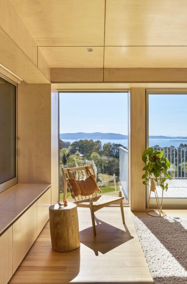 Bright, warm timber-lined interior with extensive floor-to-ceiling glazing overlooking a landscaped coastal or waterfront setting. Built-in timber cabinetry and a low media console run along the left wall, while a timber-framed lounge chair, stump side table, indoor plant, and textured rug create a calm sunlit sitting area. The room opens onto a deck with a glass balustrade, and the ceiling features flush timber panels with recessed lighting.