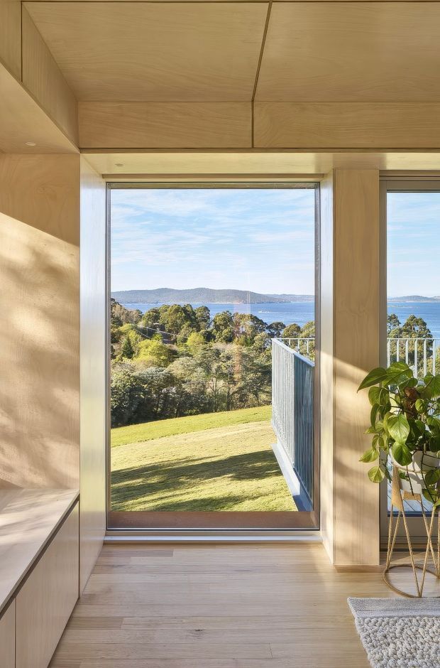 A bright, minimalist interior view centred on a large floor-to-ceiling window overlooking a landscaped sloping garden, trees, and a blue waterway with hills beyond. The room is lined with light timber cabinetry and wall/ceiling panels, with matching pale timber flooring, a potted plant, and a small textured rug. A balcony or terrace railing is visible outside.