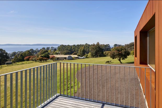 Exterior view from a raised timber-clad building, looking across a metal-balustraded deck toward a broad grassy property and a calm coastal inlet or bay. The building has clean contemporary lines, warm vertical timber cladding, large dark-framed glazing, and a simple projecting roof form. The deck is finished in pale timber, with an open landscape and water view beyond.