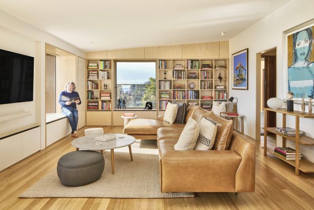 Bright, warm contemporary living room with extensive built-in timber shelving and cabinetry framing a central window, a tan leather sectional sofa, upholstered ottoman, light rug, oval coffee tables, and a wall-mounted television. The room has timber flooring, cream walls and ceiling, curated books and objects, and artwork displayed on adjacent walls.