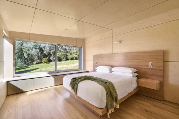A bright, minimalist bedroom with extensive warm timber wall and ceiling lining, timber flooring, and a low platform bed integrated into a full-width timber headboard. Floating bedside tables and low built-in cabinetry sit beneath large windows overlooking greenery. The room has clean contemporary detailing, concealed storage, and abundant natural light.