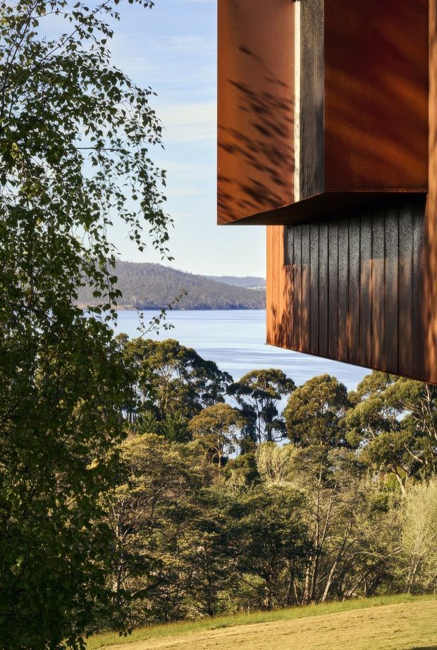 Exterior architectural detail of a contemporary building projecting over a wooded landscape, with warm rust-coloured metal cladding and dark vertically textured cladding. The structure frames a broad body of water and forested hills beyond, while trees and grassy ground fill the foreground. Strong afternoon light creates dramatic shadows across the facade.