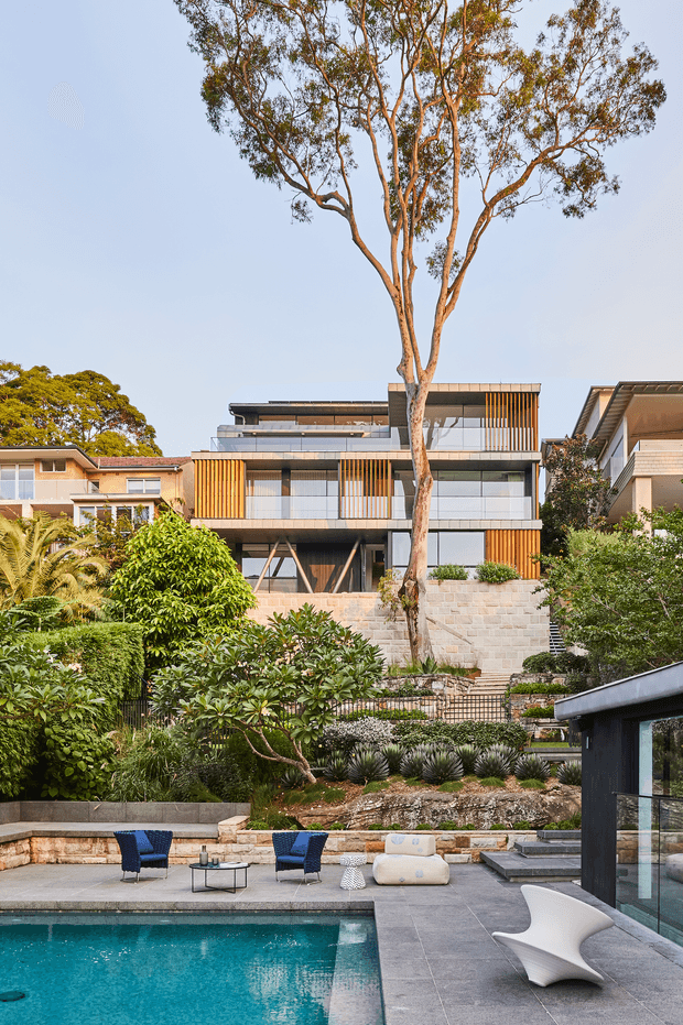 A landscaped residential property viewed from a pool terrace. The image shows a contemporary multi-level home with extensive glazing, horizontal rooflines, timber-look vertical screens, and pale masonry walls set into a sloping site. A large eucalyptus tree and dense planting occupy the foreground and middle ground, with a swimming pool, paved terrace, outdoor seating, and a dark-clad poolside structure visible at the bottom of the frame. Nearby residential buildings are visible in the background.