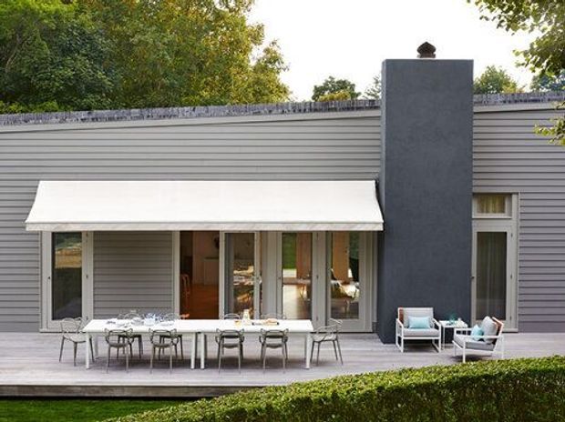 Exterior view of a low, modern residential house with a grey horizontal-clad facade, broad dark chimney, and a generous white retractable awning over a glazed outdoor dining terrace. A timber deck, dining table and chairs, lounge chairs, and clipped hedge define the outdoor entertaining area.