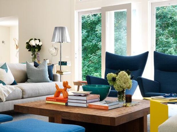 Bright, inviting living room with a large timber coffee table, light upholstered sofa, deep navy lounge chairs, blue accents, books, decorative objects, and flowers. Tall white-framed windows bring in abundant natural light and overlook greenery.