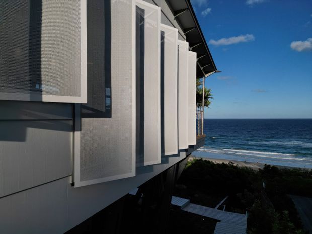 Exterior architectural view of a contemporary multi-level building overlooking the ocean. The facade features projecting white rectangular perforated or screened panels, dark horizontal cladding, and a sharply angled roof edge. The building appears elevated above a landscaped coastal site, with expansive ocean and beach visible beyond.