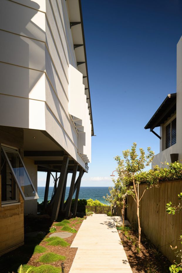 A sunlit coastal residential external walkway descends between two modern houses toward a visible ocean horizon. The left building has white horizontal-panel cladding, projecting geometric balconies or fins, dark structural supports, and deep roof overhangs. The right house has dark roof trim, light walls, and a tall timber privacy fence with landscaped planting. A pale timber boardwalk and stepping stones lead toward the water beneath a clear blue sky.
