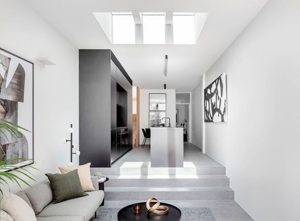 A bright, narrow contemporary open-plan interior viewed from a lower living area toward a compact kitchen and dining zone. The space features a grey sectional sofa, dark built-in cabinetry, a central island, pale grey tiled flooring, white walls, a large skylight, minimalist linear lighting, and several steps creating a split-level arrangement. Abstract artwork and indoor planting add visual interest.