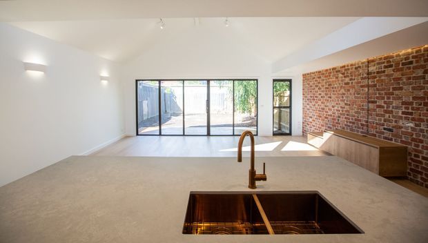 Bright open-plan kitchen interior viewed from behind a large pale stone island benchtop. The room features a deep undermount double sink with a curved brushed metal tap, dark timber cabinetry, exposed red brick feature wall, white walls and ceiling, polished concrete or light-toned flooring, and extensive black-framed glazing opening to an outdoor courtyard or patio.