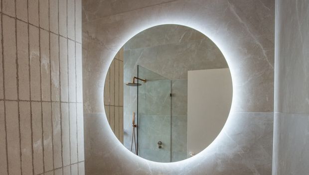 Close-up of a round backlit mirror mounted on a tiled bathroom wall. The mirror reflects a frameless glass shower enclosure with brushed metal fittings, while large pale stone-look tiles create a softly textured, warm neutral finish.
