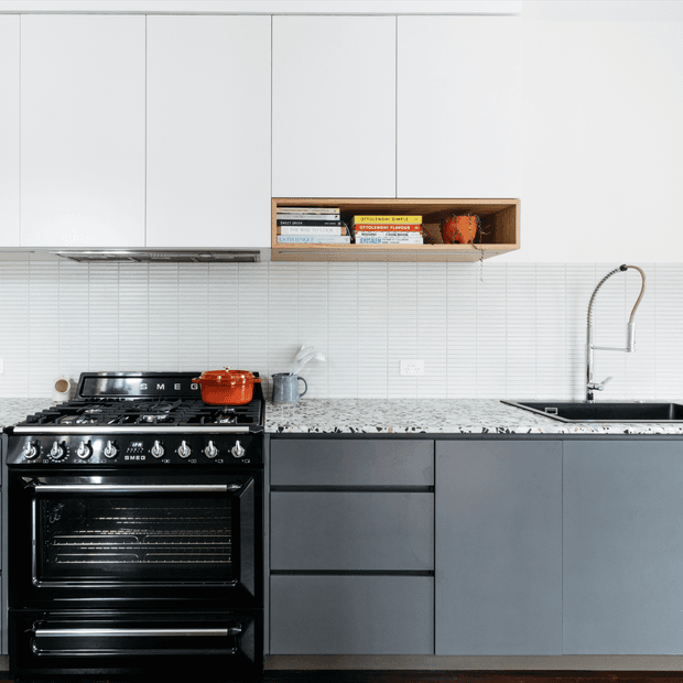 A contemporary kitchen with a black freestanding range cooker, dark grey lower cabinetry, white upper cabinets, a white-and-grey speckled stone benchtop, and a fine white tiled splashback. An open timber shelf displays books and pottery, while a stainless steel gooseneck tap and undermount sink sit to the right.