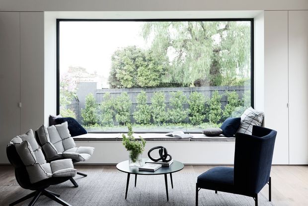A refined contemporary living room centred around a large black-framed picture window overlooking a landscaped garden. Built-in white cabinetry and a deep window seat frame the glazing, while a grey upholstered lounge chair, dark navy armchair, round glass coffee table, and textured neutral rug create a calm, minimalist seating area.