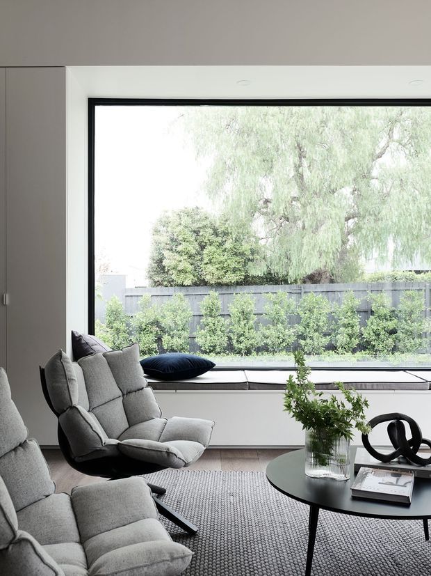 A bright, minimalist living room centered on a large floor-to-ceiling black-framed window overlooking a planted residential garden. Two sculptural grey upholstered lounge chairs sit on a textured grey rug, with a low round black coffee table holding a glass vase and decorative objects. Built-in grey cabinetry lines the left wall, and a slim window seat runs beneath the glazing.