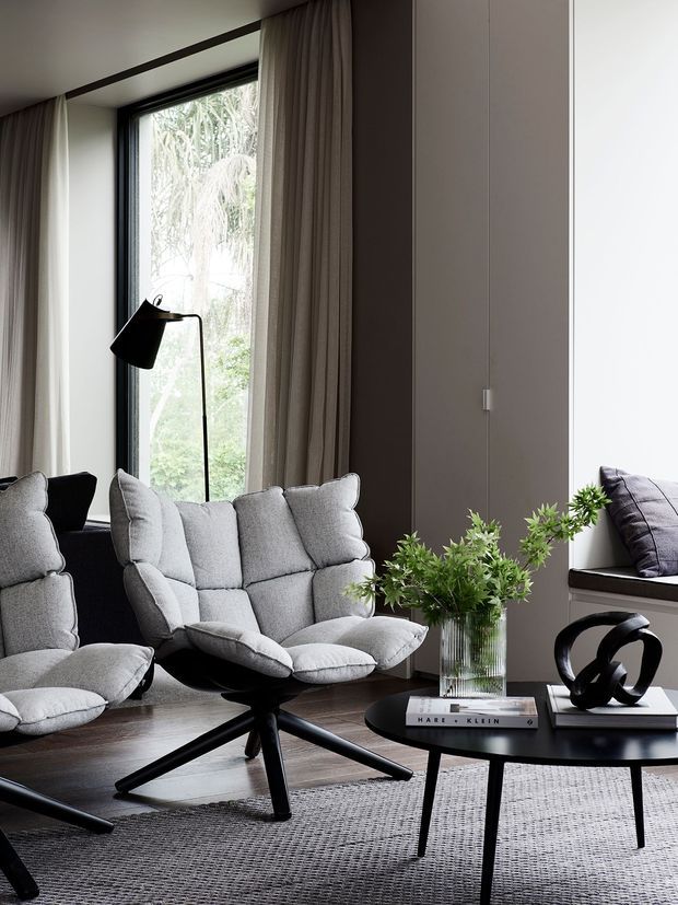 A refined contemporary living room with two plush grey lounge chairs arranged around a low black coffee table. Floor-to-ceiling windows with dark frames and full-length taupe curtains overlook greenery, while built-in wall cabinetry and a window seat line the opposite side. A black sculptural floor lamp, glass vase with greenery, decorative objects, and a textured grey rug complete the calm, modern setting.