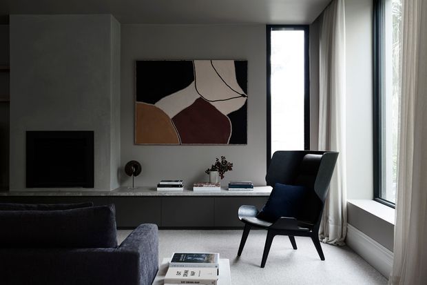 A refined contemporary living room with a dark upholstered sofa in the foreground, a sculptural black lounge chair beside full-height corner glazing, and a long minimalist built-in media console beneath abstract artwork. The room has soft grey walls, pale carpet, floor-to-ceiling curtains, and restrained monochromatic furnishings with warm muted accents.