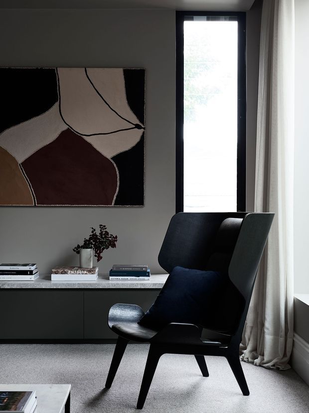 A refined, moody reading corner in a contemporary interior. A sculptural black lounge chair with a dark cushion sits beside a tall narrow window dressed with full-length sheer curtains. Behind it is a dark built-in sideboard with a pale stone-look benchtop, books, and a small plant, while a large abstract neutral-toned artwork hangs on the grey wall. The room has a low-contrast, monochromatic palette with tactile finishes and soft natural light.