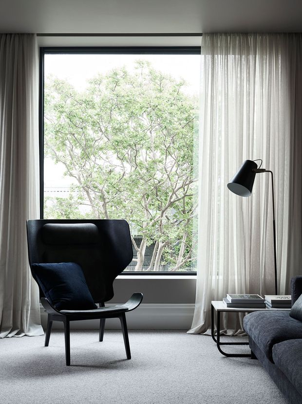 A refined living room corner with a sculptural black lounge chair, dark upholstered sofa, slim black floor lamp, and low side table with books. Full-height sheer curtains frame a large black-framed window overlooking leafy trees, while pale carpet and neutral walls create a calm, softly lit atmosphere.