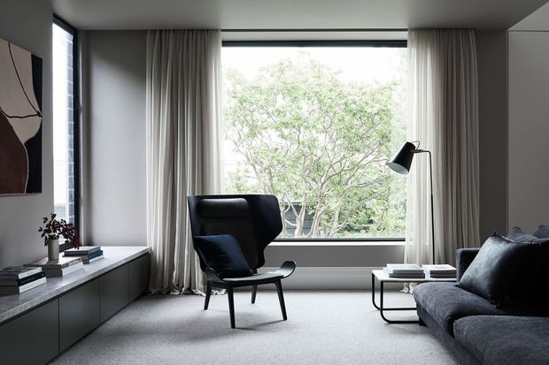 A refined contemporary living room with a low built-in media cabinet, textured grey carpet, a dark sculptural lounge chair, charcoal sectional sofa, black coffee tables, and full-height curtains framing a large picture window overlooking trees. The room has a restrained monochromatic palette with soft natural light and minimalist furnishings.